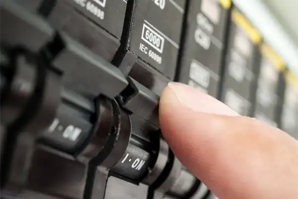 Close-up of a hand turning on a circuit breaker switch, showing the label "6000" and "IEC 60898" on the device.