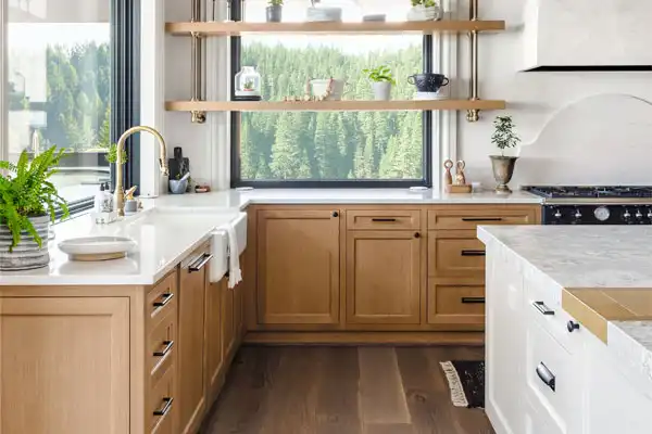A modern kitchen showcases wooden cabinets, white countertops, and a large window with views of trees. Open shelves display plants and dishes. To the left is a sink with a gold faucet, ready for any plumbing repair needs by a nearby plumber.