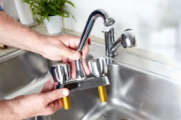Person holding a detached kitchen faucet above a stainless steel sink, with two handles and exposed brass threads.