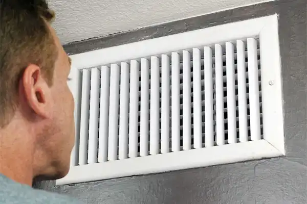 A person examining a wall-mounted air vent with white slats.