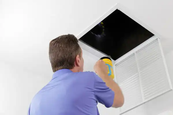 A person in a blue shirt shines a flashlight into a dark attic or ventilation opening in the ceiling.