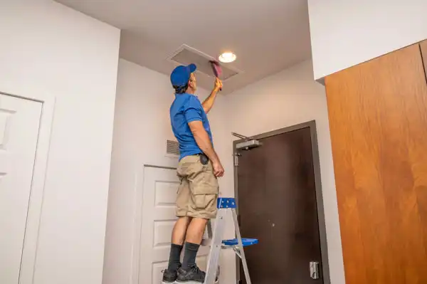 Person in a blue shirt and cap standing on a ladder, cleaning a ceiling vent with a brush in a room.