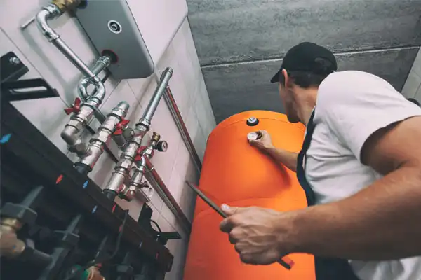 Technician in a cap and uniform checking a large orange boiler tank, holding a wrench, with various pipes visible on the wall.