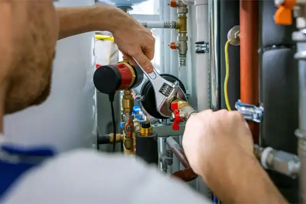 Person using a wrench to tighten a valve on a network of pipes and valves in a mechanical room.