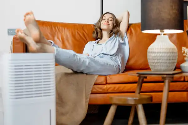 Woman relaxing on a brown sofa, with a blanket, feet up on a table. A dehumidifier and lamp are in the foreground.