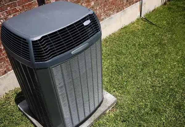 An outdoor air conditioner unit sits on a concrete pad beside a brick wall, surrounded by grass.