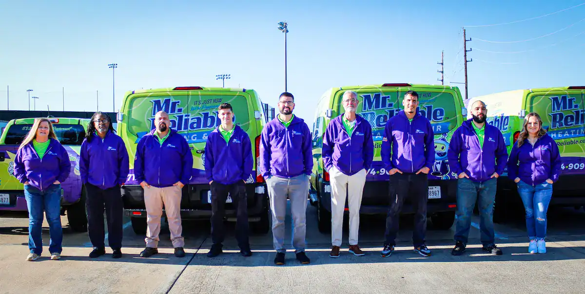 Eight people wearing purple jackets stand in front of green service vans parked in a lot under a clear blue sky.