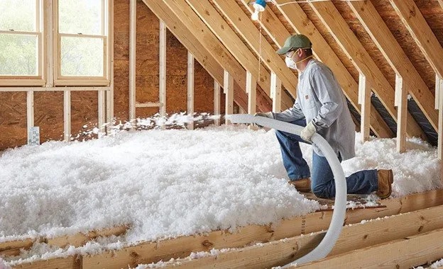 A person kneels in an unfinished attic, wearing a mask and cap, while performing Blown-In Insulation Installation to improve energy efficiency and support air conditioning by spraying loose-fill material onto the floor between wooden beams.